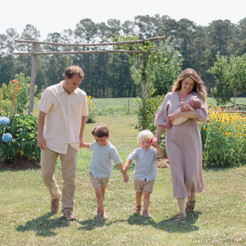 family walking through flower garden. mom holding newborn girl, 2 young boys in the middle and dad on other side all holding hands in a line.